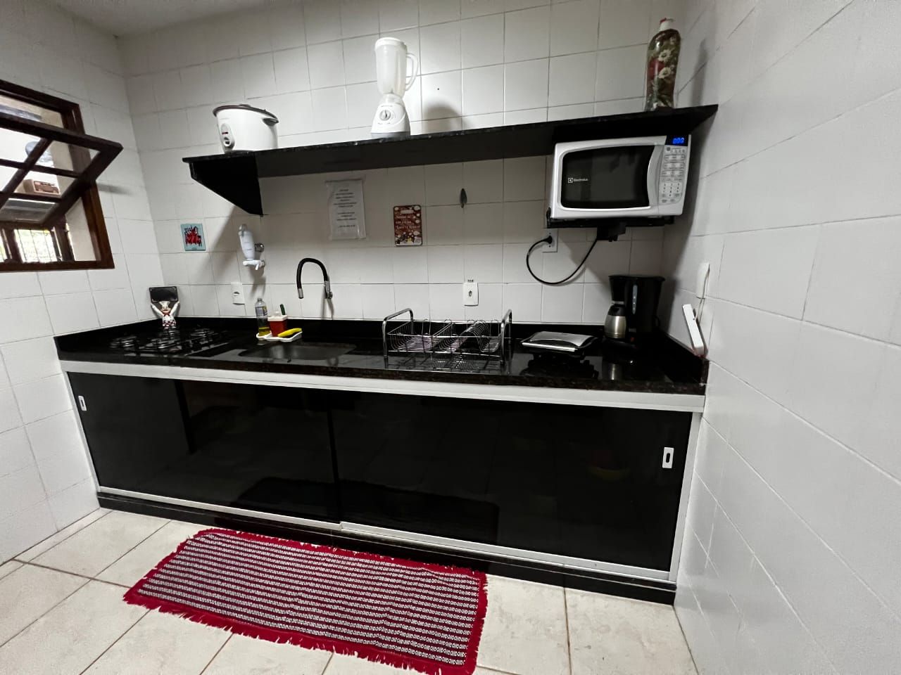 A kitchen with a black countertop, white tiles, a microwave, a blender, and a red rug.