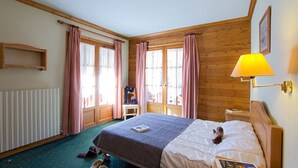 Ein gemütliches Hotelzimmer mit Holzbalkendecke, einem Bett mit weißer Bettdecke, einem kleinen Nachttisch, einer Lampe und einem Fenster mit karierten Vorhängen.