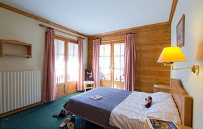 A cozy hotel room with a wooden ceiling, a bed with a white comforter, a small bedside table, a lamp, and a window with checkered curtains. - À 50m des remontées | T3 central & coin tranquille (Les Deux Alpes)