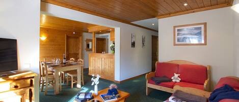 A cozy room with a wooden ceiling, a dining area with a wooden table and chairs, a red sofa, and a television.