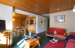 A cozy room with a wooden ceiling, a dining area with a wooden table and chairs, a red sofa, and a television. - À 50m des remontées | T3 central & coin tranquille (Les Deux Alpes)