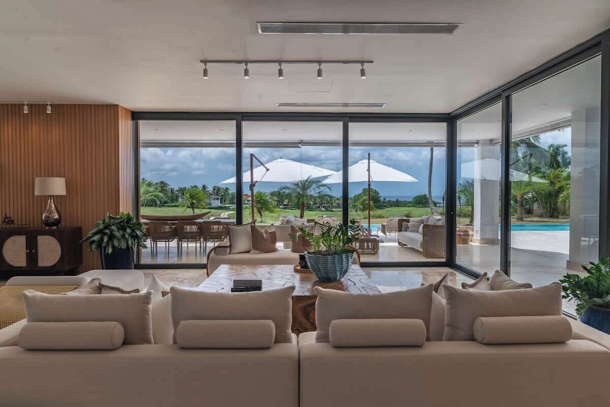 A modern living room with a large glass wall, a sofa, a coffee table, and a view of a pool and garden.