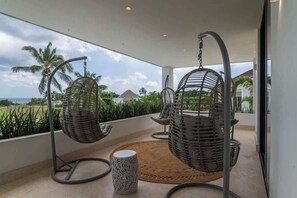 A balcony with wicker chairs and a woven rug, overlooking a landscape with palm trees and a thatched-roof building. - Villa Cinco Ríos · Infinity Pool, Golf & Ocean Views, Staff Included (La Romana)