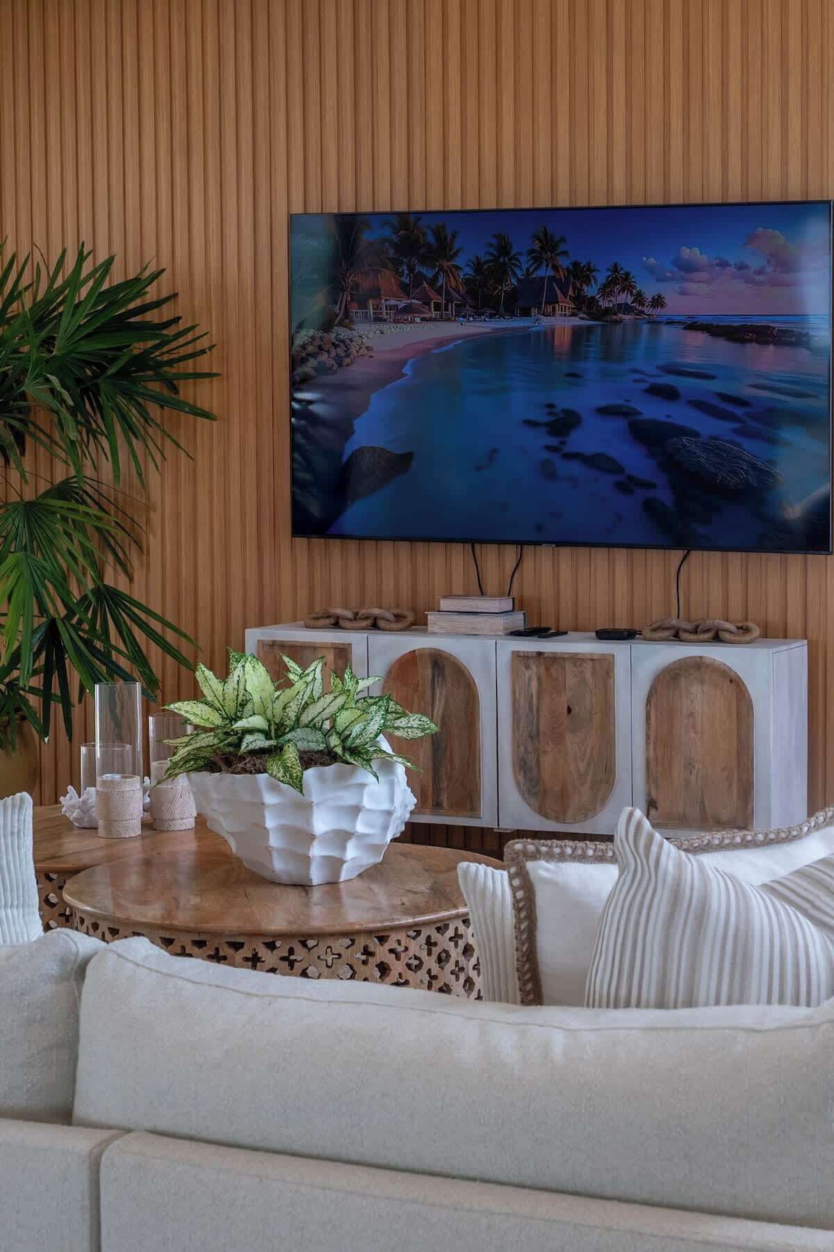 Ein Wohnzimmer mit einem großen Flachbildfernseher an einer Holzwand, einem runden Holztisch mit einem Blumentopf und einem weißen Sofa.