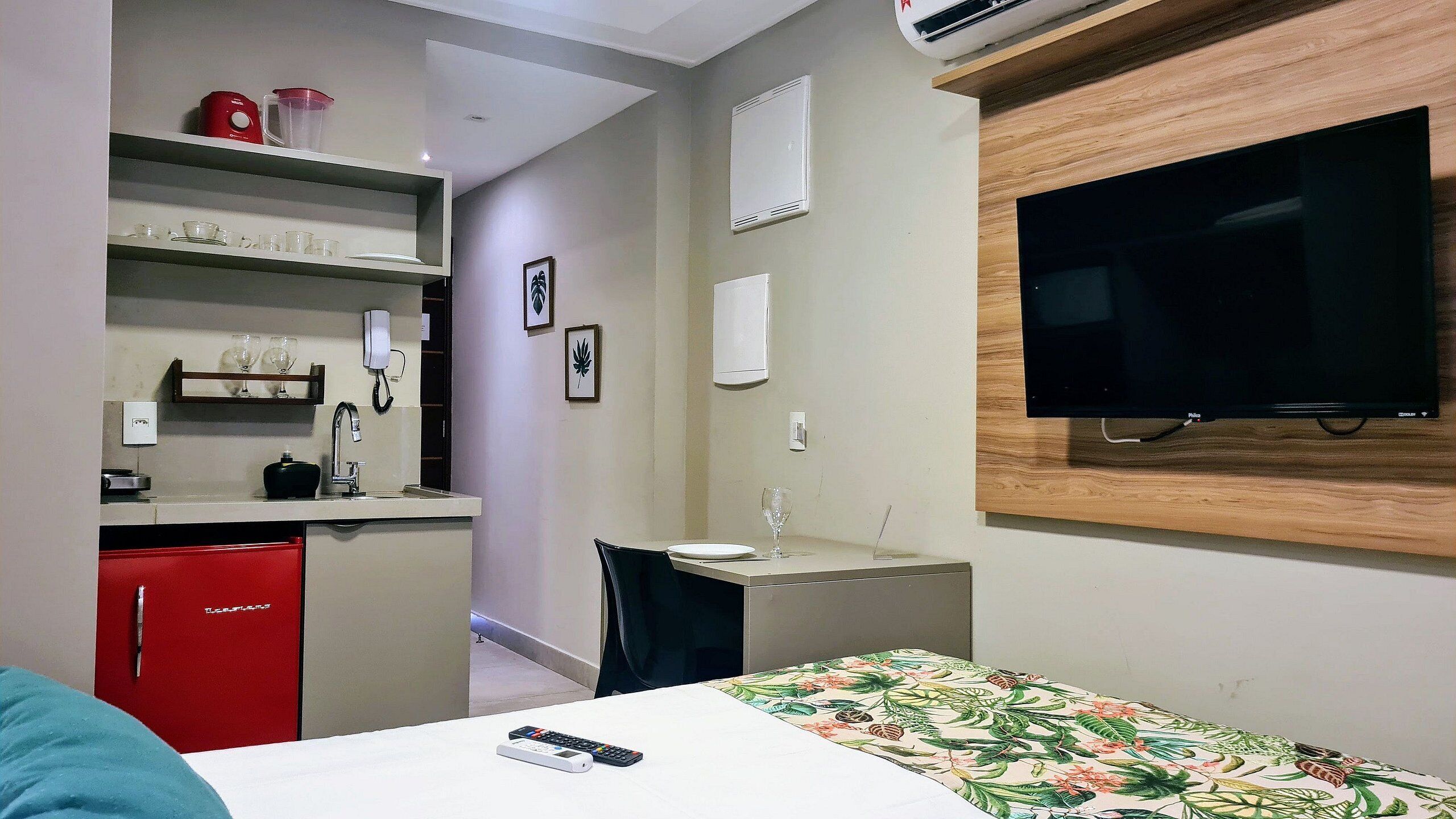A compact hotel room with a bed, a desk with a chair, a red minibar, a television, and a bathroom with a shower.