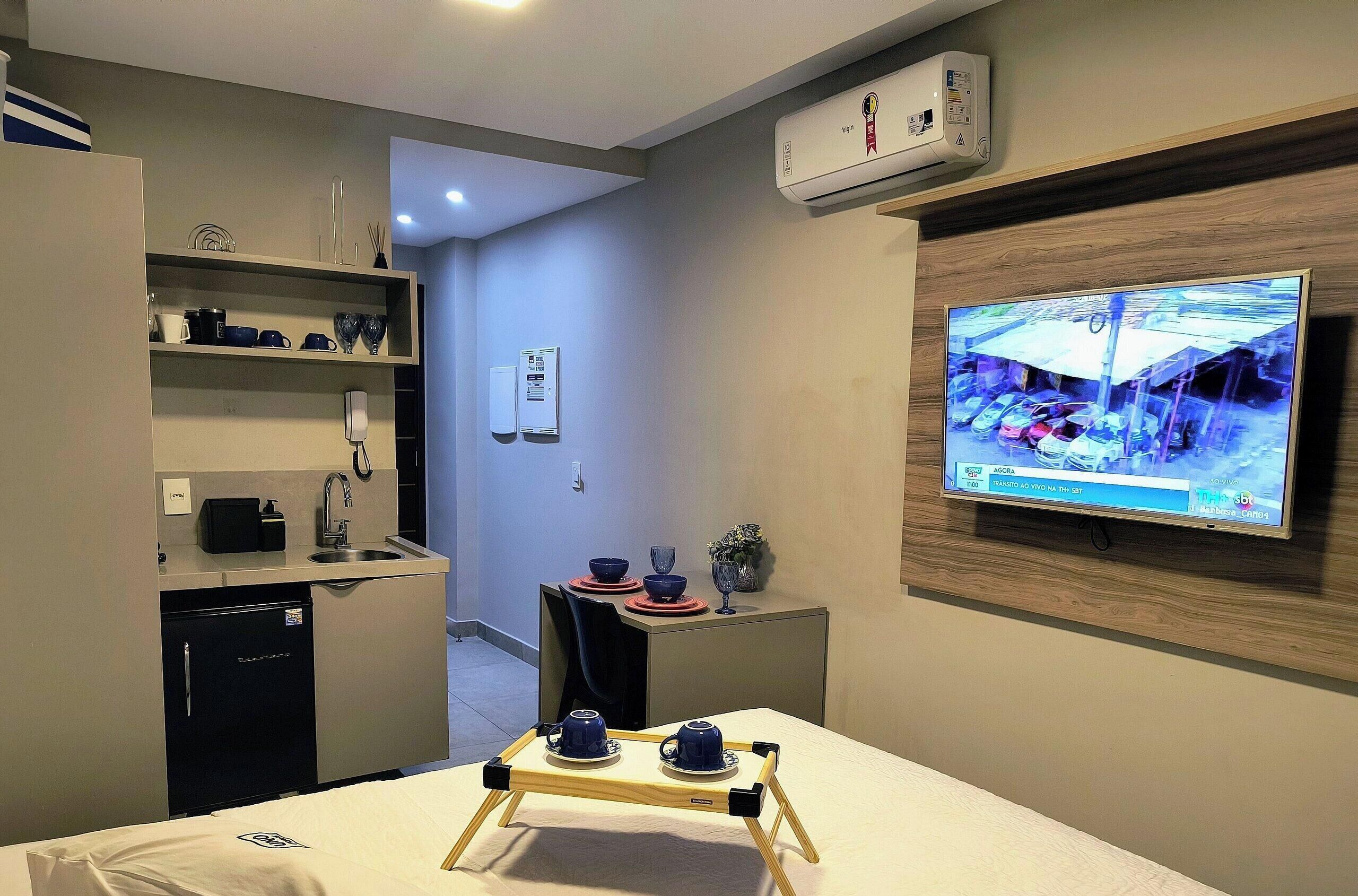 Habitació d'hotel amb televisió penjada a la paret, cuineta, zona de menjador amb taula per a dues persones i llit amb mantell blanc.