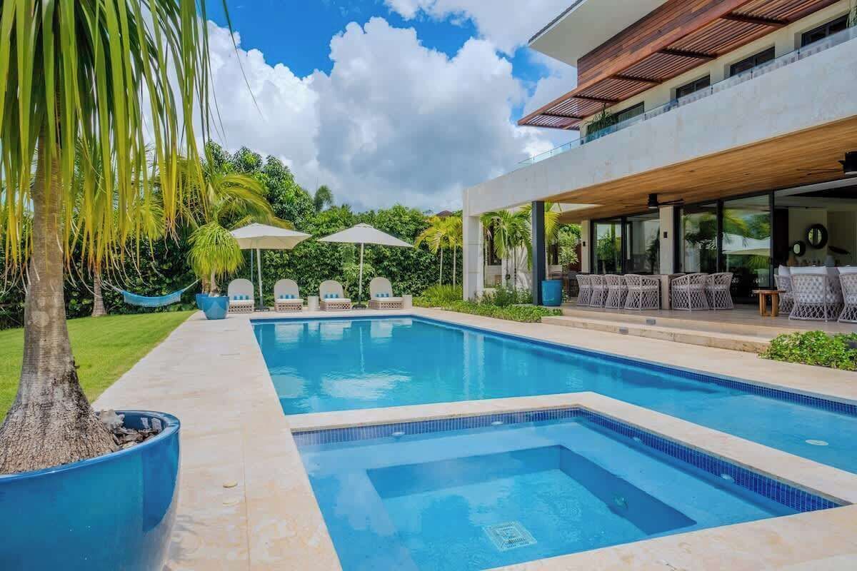 A swimming pool with lounge chairs and umbrellas, surrounded by greenery and a modern building.