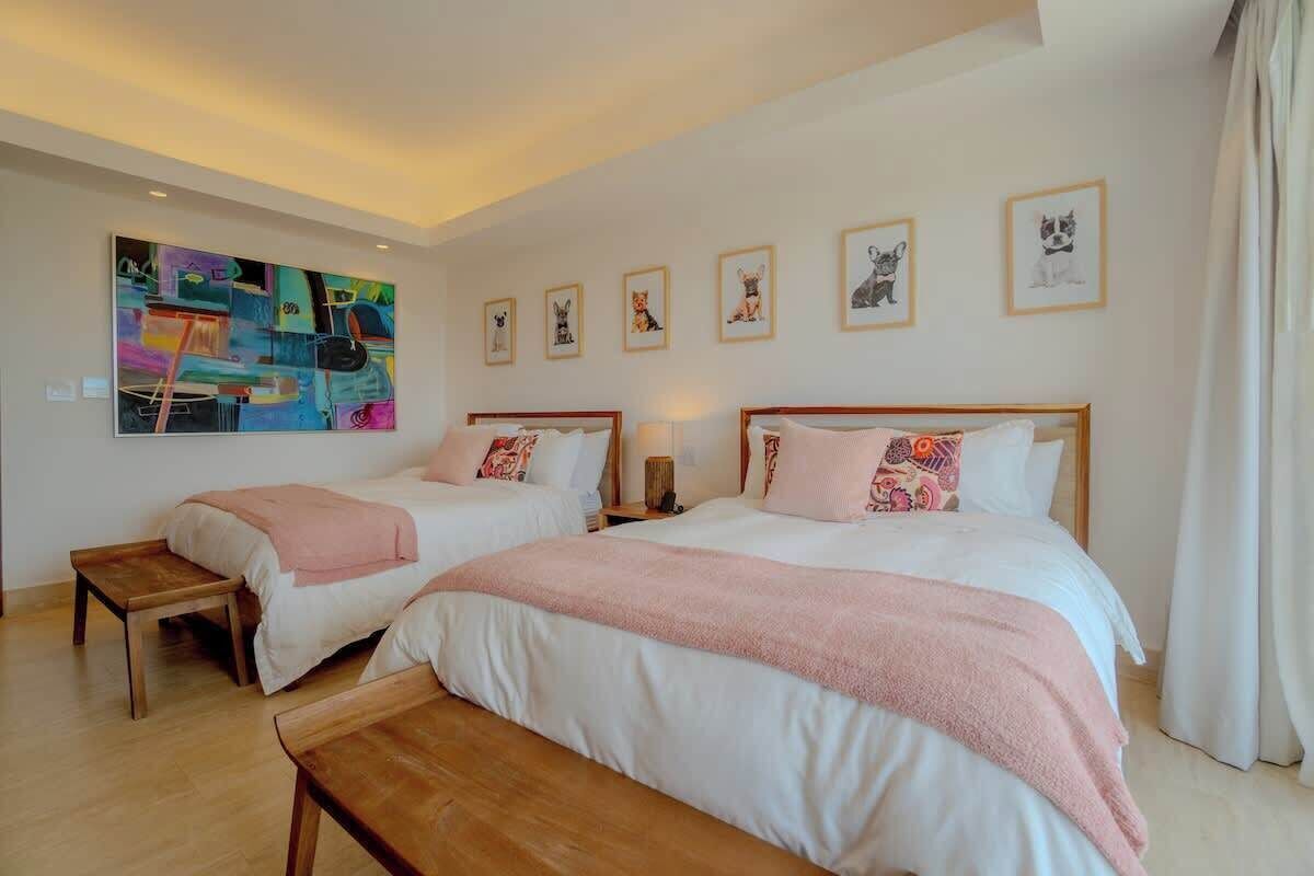 A hotel room with two beds, a bench, a painting, and framed pictures on the wall.