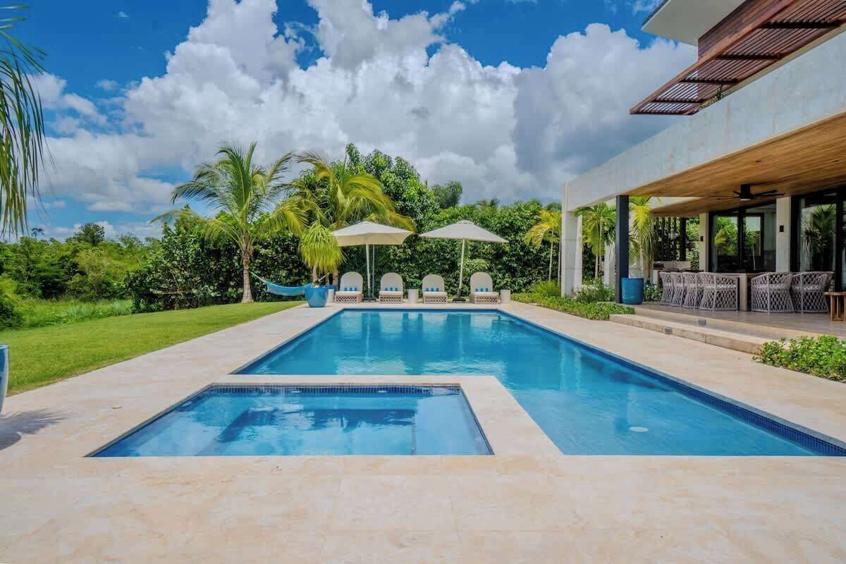 A rectangular swimming pool with a tiled deck, surrounded by lush greenery and palm trees.
