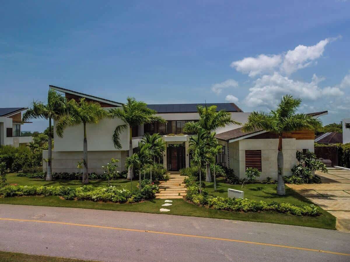 A modern two-story house with a paved driveway, a stone pathway leading to the entrance, and a well-maintained lawn with palm trees.