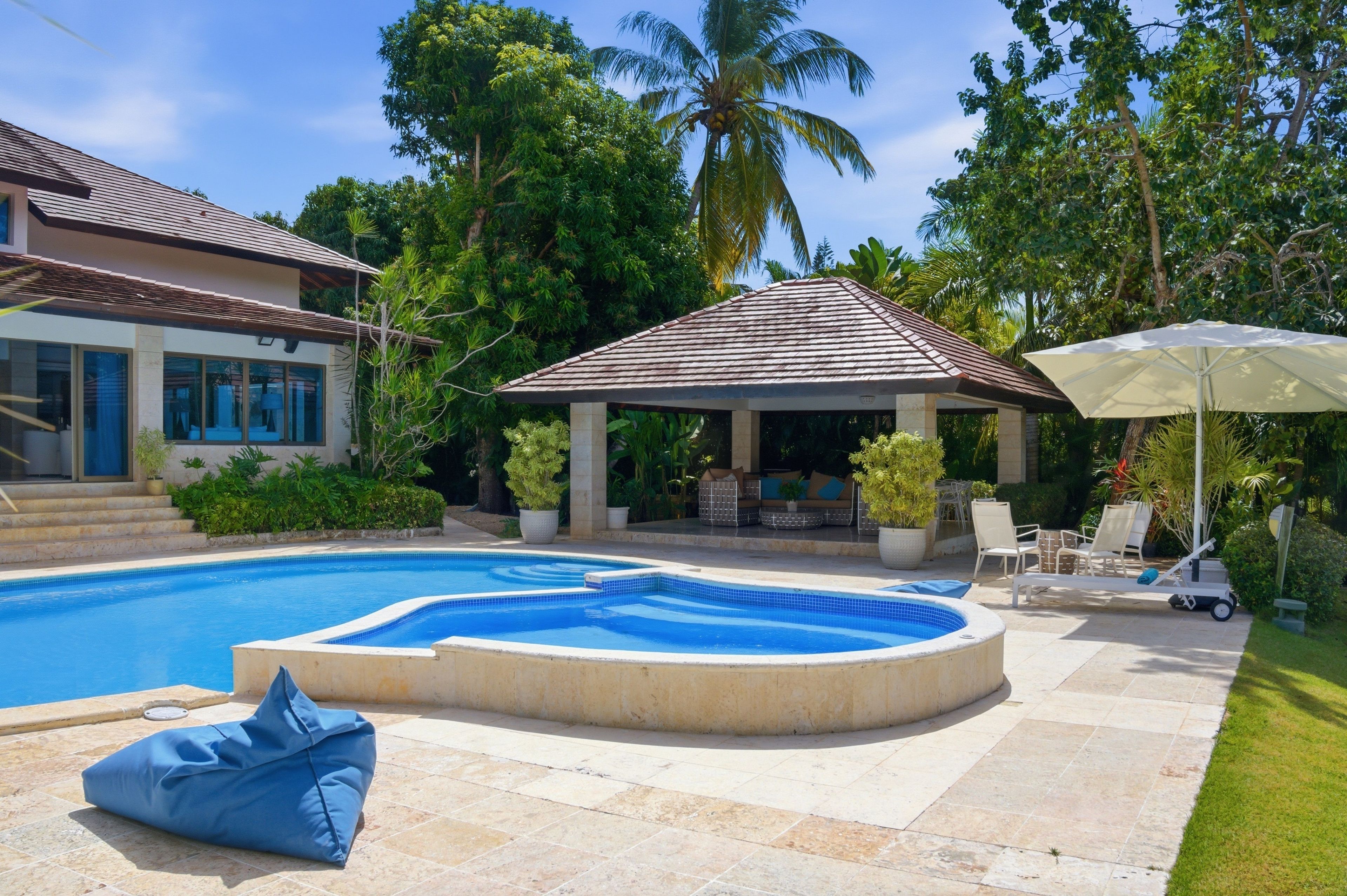A swimming pool with a hot tub, surrounded by a covered seating area, palm trees, and a house with a tiled roof.