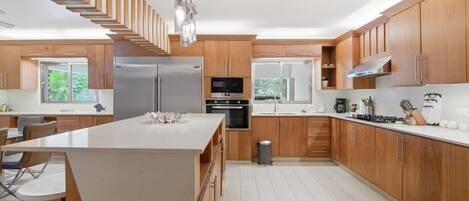 A modern kitchen with wooden cabinetry, a central island, stainless steel appliances, and a view of greenery outside.