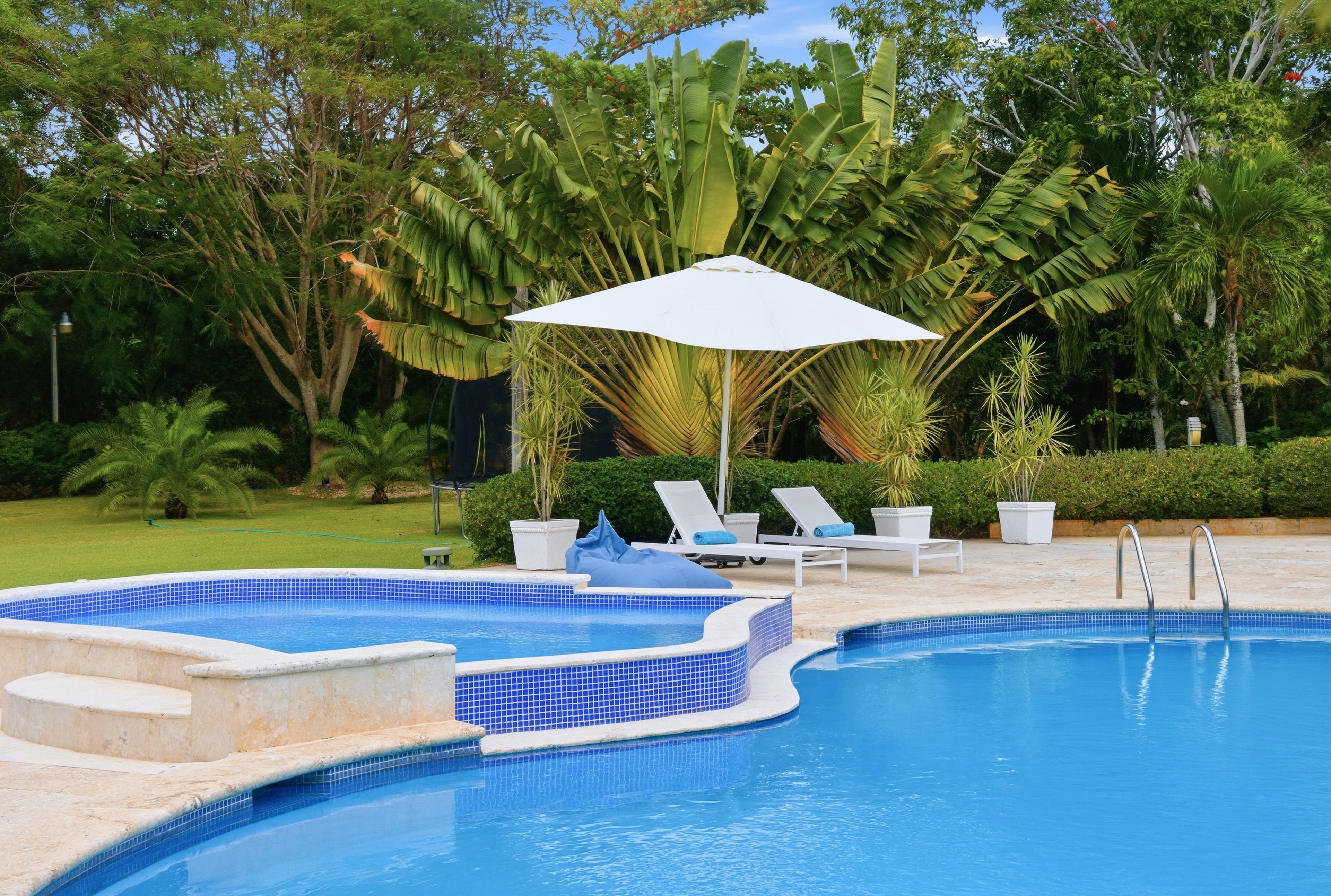 A swimming pool with a white umbrella, surrounded by lounge chairs and tropical plants.