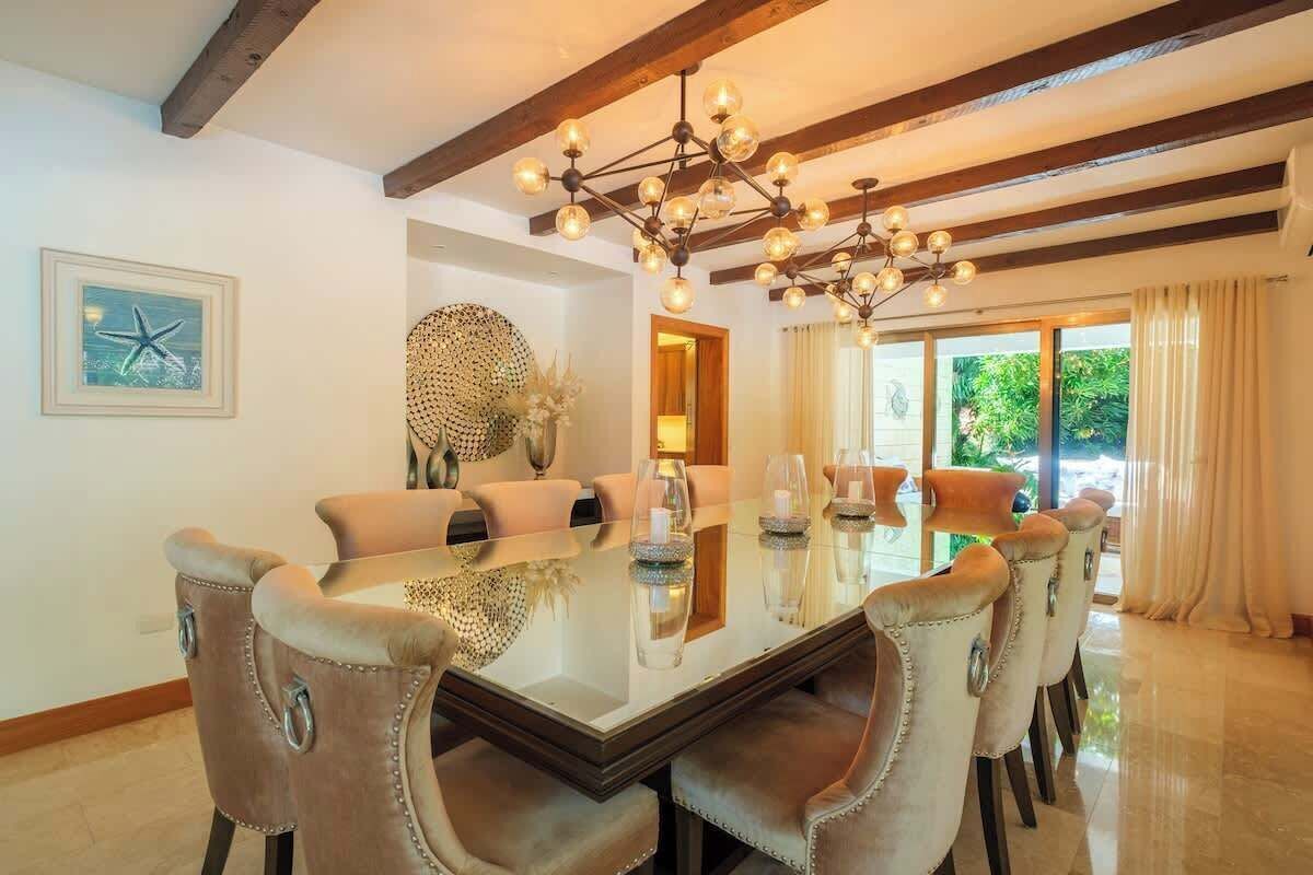 Un espace repas avec une table en verre, des chaises rembourrées, un lustre et une vue sur la verdure environnante.