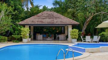 A swimming pool with a tiled deck, a covered seating area with chairs and a table, surrounded by lush greenery.