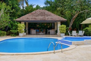 A swimming pool with a tiled deck, a covered seating area with chairs and a table, surrounded by lush greenery.