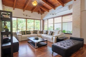 A spacious living room with a high ceiling, large windows, a sectional sofa, a coffee table, and a black ottoman.