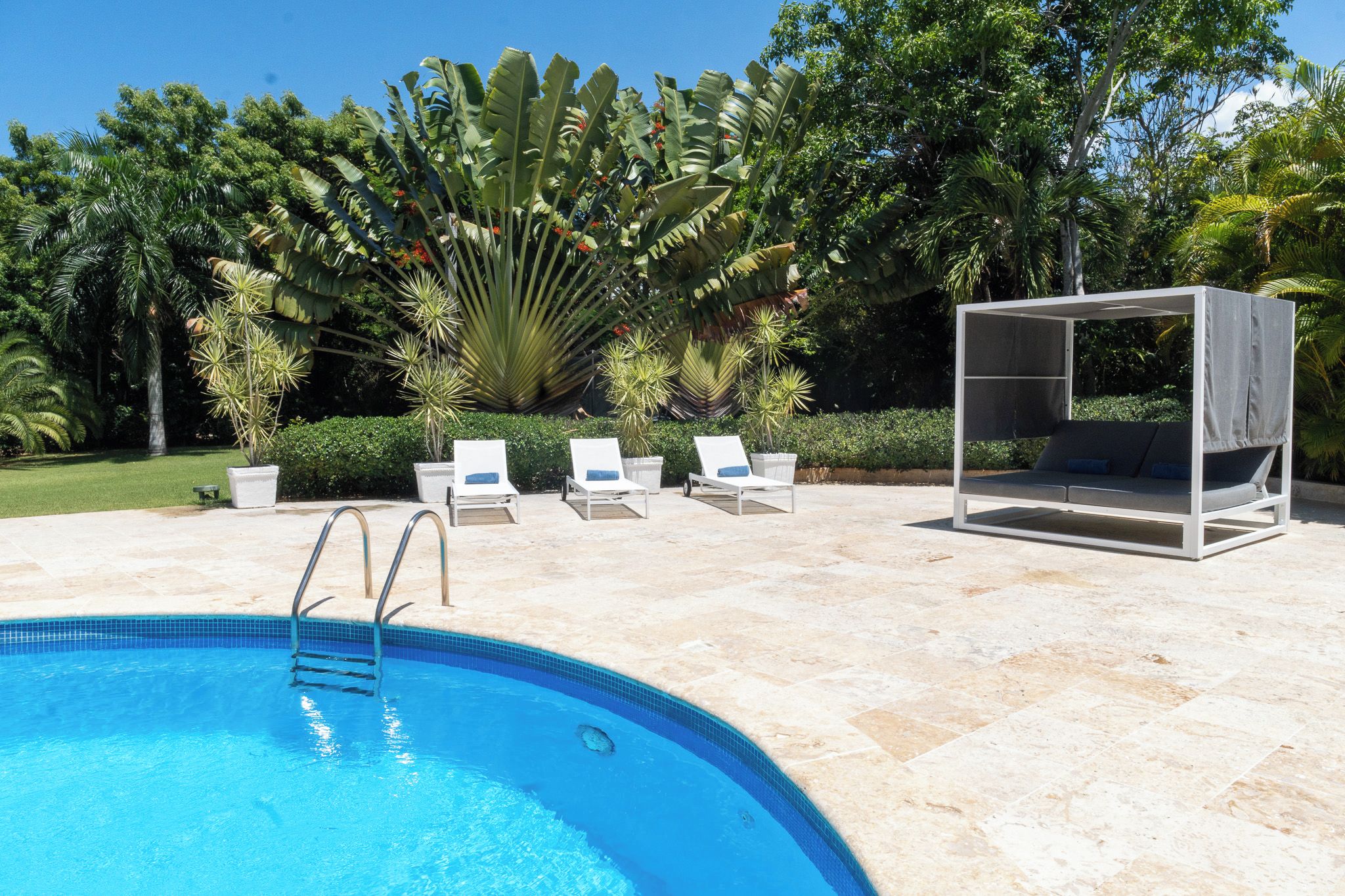 A swimming pool with a clear blue water, surrounded by a tiled deck. There are lounge chairs and a shaded seating area with a canopy.