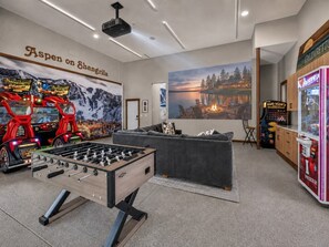 A room with a foosball table, a sofa, a wall with a scenic painting, and a claw machine.