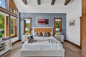 A bedroom with a large bed, two bedside tables, a headboard, and a view of the outdoors through large windows.