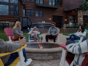 A group of people seated around a fire pit in an outdoor setting with a wooden cabin in the background.