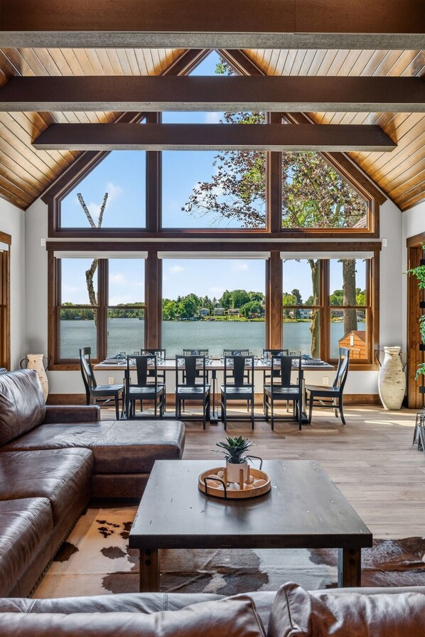 A spacious room with a large window overlooking a lake, furnished with a leather sofa, dining table, and chairs.