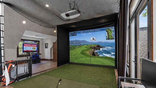 Een kamer met een groot scherm waarop een golfbaan te zien is, een bureau met een toetsenbord, en een golfbag.