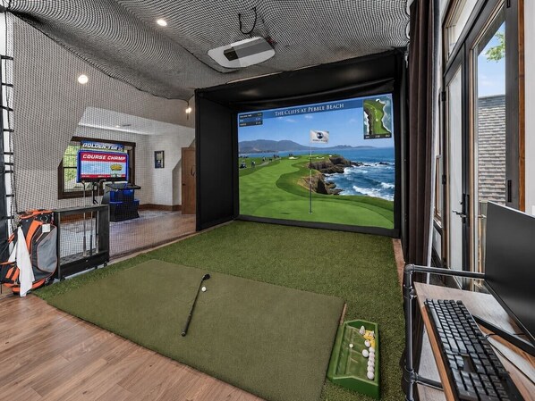 A room with a large screen displaying a golf course, a desk with a keyboard, and a golf bag.