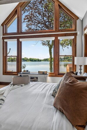 A bedroom with a large window overlooking a lake, a bed with pillows, a bedside table, and a lamp.