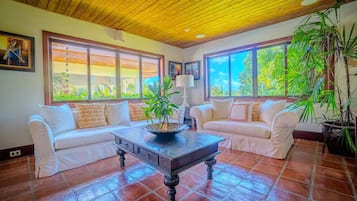 A room with two white sofas, a wooden coffee table, and large windows with a view of greenery.
