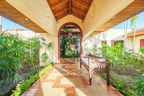 Un paseo techado con un banco de madera, rodeado de abundante vegetación y suelo de baldosas.