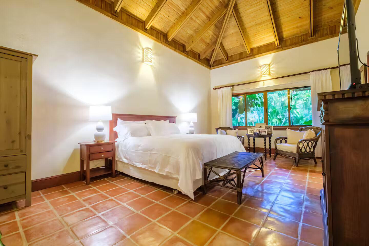 A bedroom with a bed, bedside tables, a chair, a desk, and a window with a view of greenery.