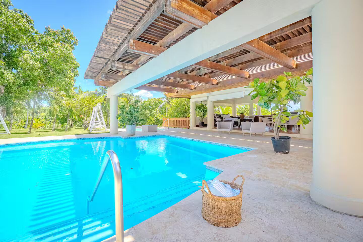 A swimming pool with a wooden pergola overhead, surrounded by greenery and outdoor furniture.