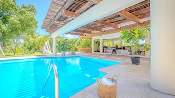 A swimming pool with a wooden pergola overhead, surrounded by greenery and outdoor furniture.
