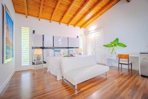 Une pièce spacieuse avec un plafond en bois, un lit avec une literie blanche, un bureau avec une chaise et une grande plante verte.