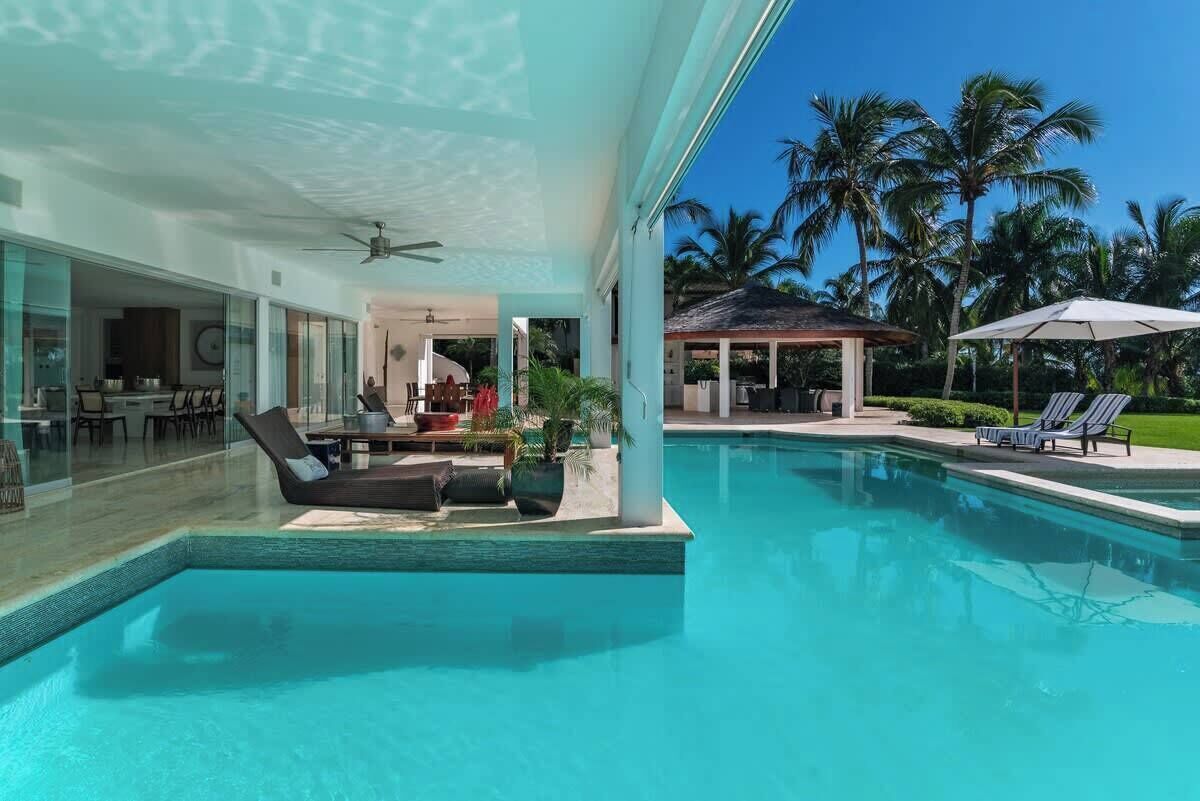 A swimming pool with lounge chairs, a covered seating area, and a view of palm trees.
