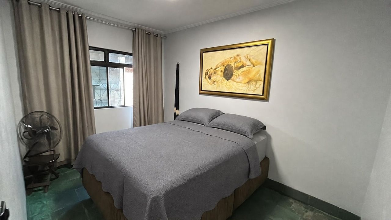 Un dormitorio con una cama, un ventilador, una ventana con cortinas y un cuadro en la pared.