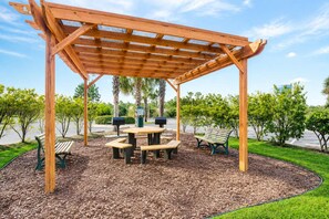 Eine hölzerne Pergola mit Bänken und Picknicktischen in einer angelegten Grünfläche.