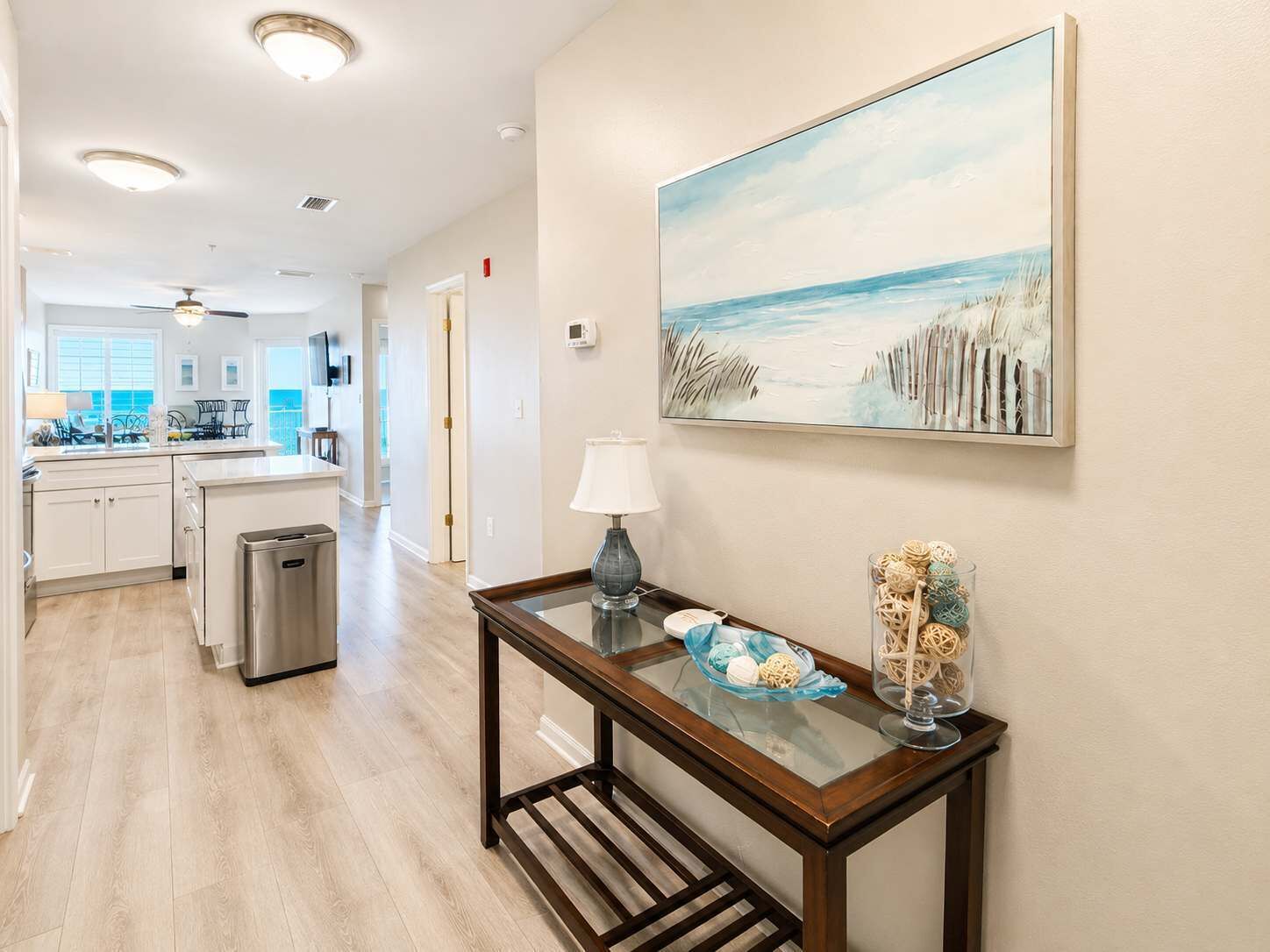 A hallway with a dining table, a lamp, and a painting of a beach scene.