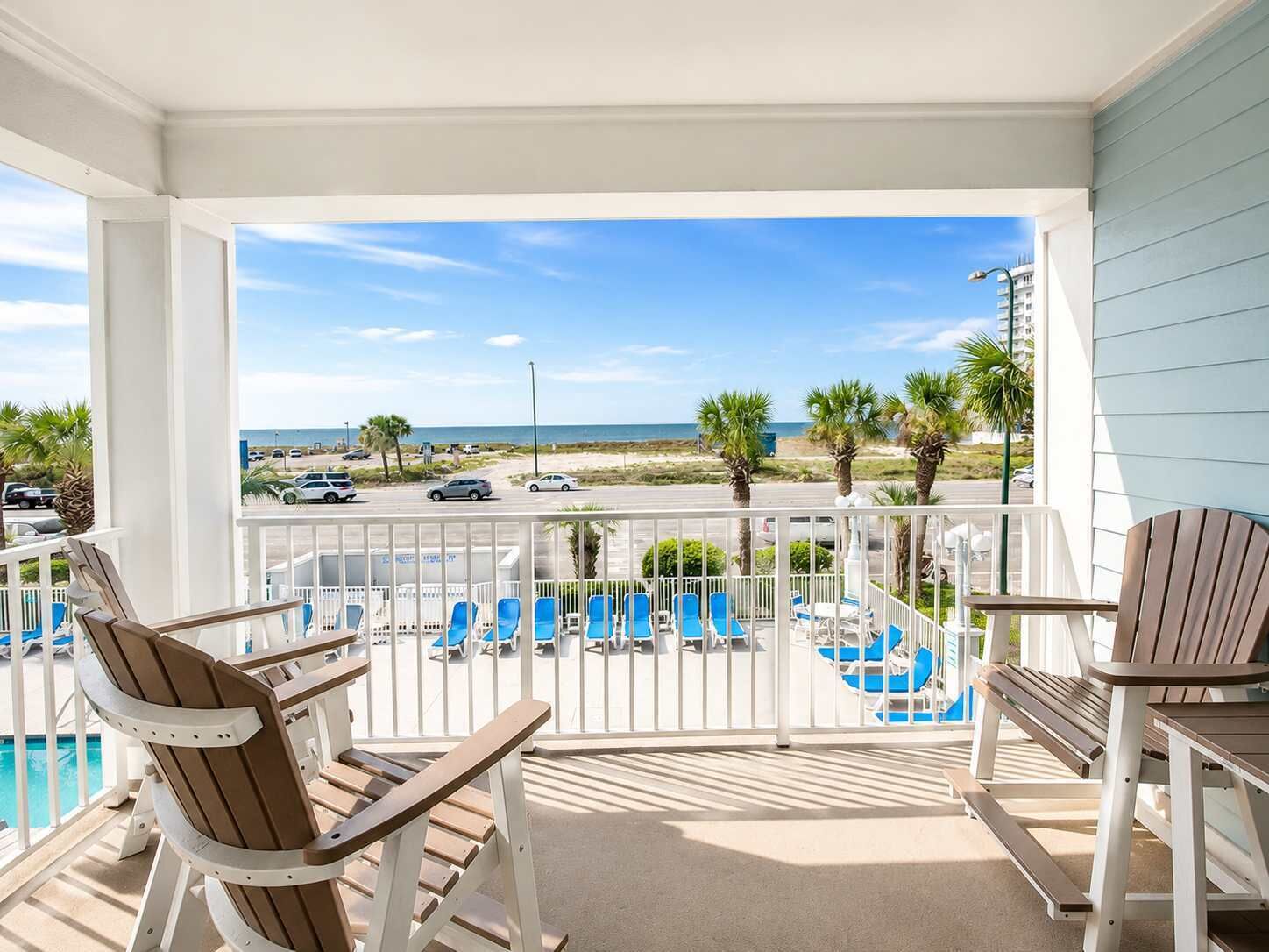 A balcony with a view of the beach, poolside chairs, and a table.