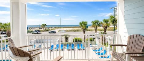 A balcony with a view of the beach, poolside chairs, and a table.