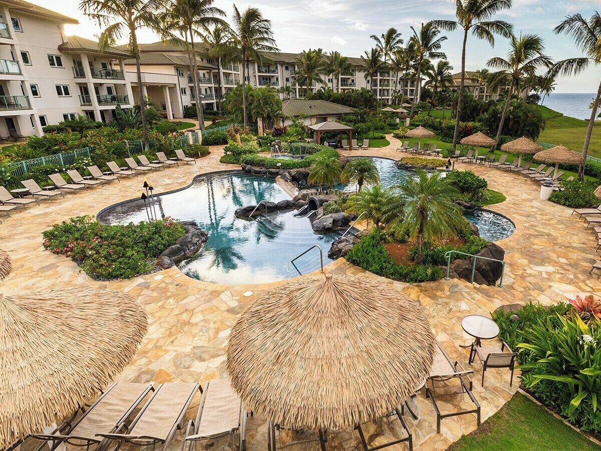 Ein Resort mit einem runden Pool, umgeben von Liegestühlen und Sonnenschirmen, Palmen und einem Gebäude im Hintergrund.