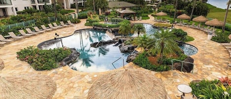 Un resort con una piscina circular rodeada de sillones de descanso y sombrillas, palmeras y un edificio al fondo.