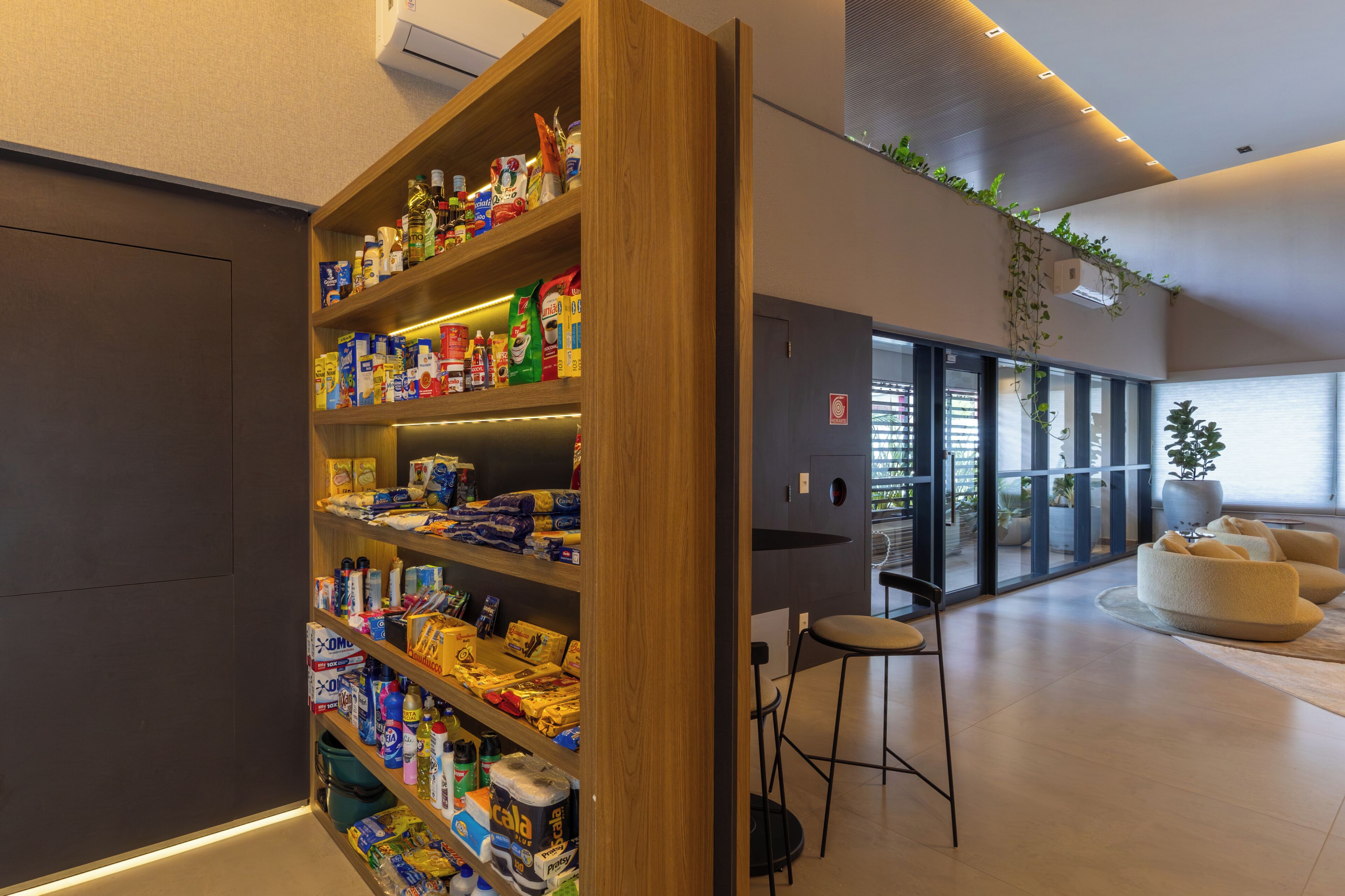 Un intérieur moderne avec une étagère en bois remplie de divers produits alimentaires, un coin salon avec une table ronde et une vue sur l’extérieur grâce à de grandes fenêtres.