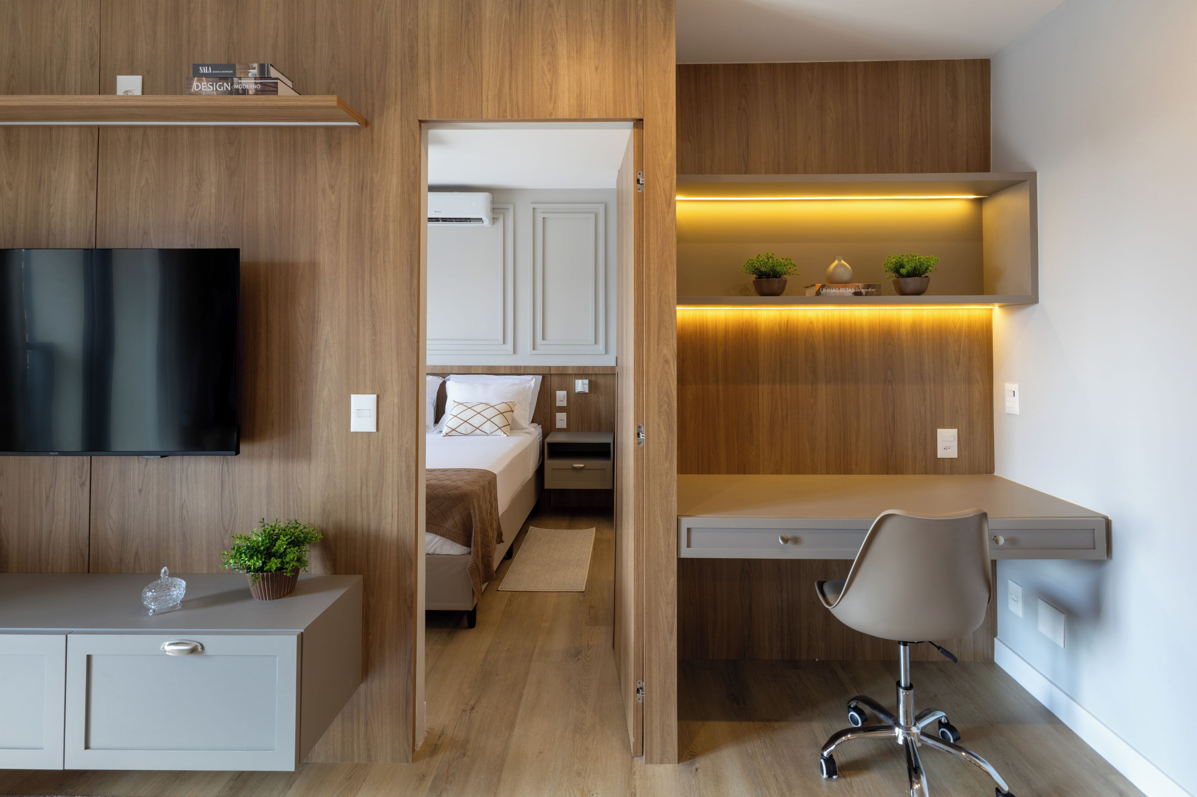 Una habitación de hotel moderna con interior de madera, un televisor de pantalla plana, un escritorio con una silla, una cama con cabecera blanca y una planta pequeña sobre el escritorio.