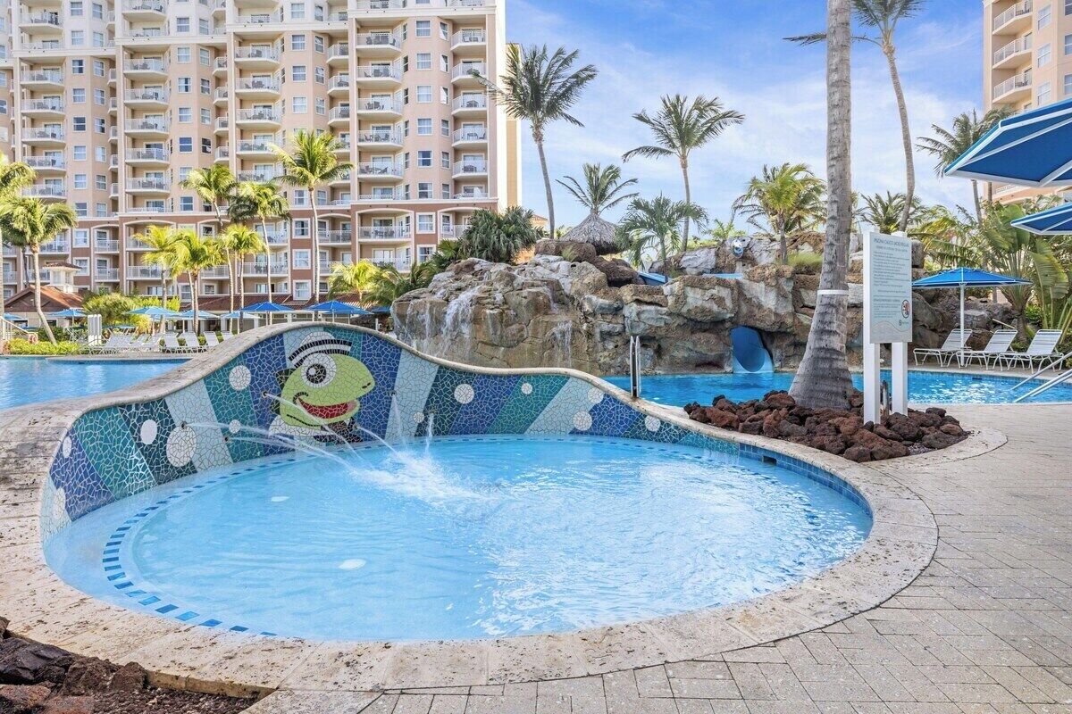 Ein Schwimmbecken mit Mosaikverziertem Rand, umgeben von Palmen und einem Hotel im Hintergrund.