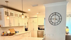 A modern kitchen with white cabinetry, a central island with bar stools, pendant lighting, and a wall-mounted clock. - Spacious 5-bedroom Modern Retreat with Heated Pool Near Beach and Cove Shopping (Deerfield Beach)