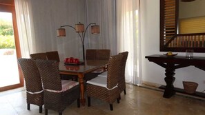 A dining area with a wooden table and wicker chairs, a small side table with a basket, and a view of greenery outside. - Caribbean bungallow with 2 bedrooms in Cap Cana (Punta Cana)