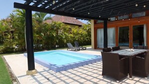 Ein Pool mit Liegestühlen und einem Tisch unter einer Pergola, umgeben von Palmen und einem Gebäude mit großen Fenstern.