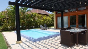 A swimming pool with lounge chairs and a table under a pergola, surrounded by palm trees and a building with large windows. - Caribbean bungallow with 2 bedrooms in Cap Cana (Punta Cana)